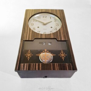 May include: A vintage Lorus clock with a wooden frame and a round clock face. The clock face has black numerals and hands. Below the clock face is a display showing the date and day. The clock has decorative gold accents.