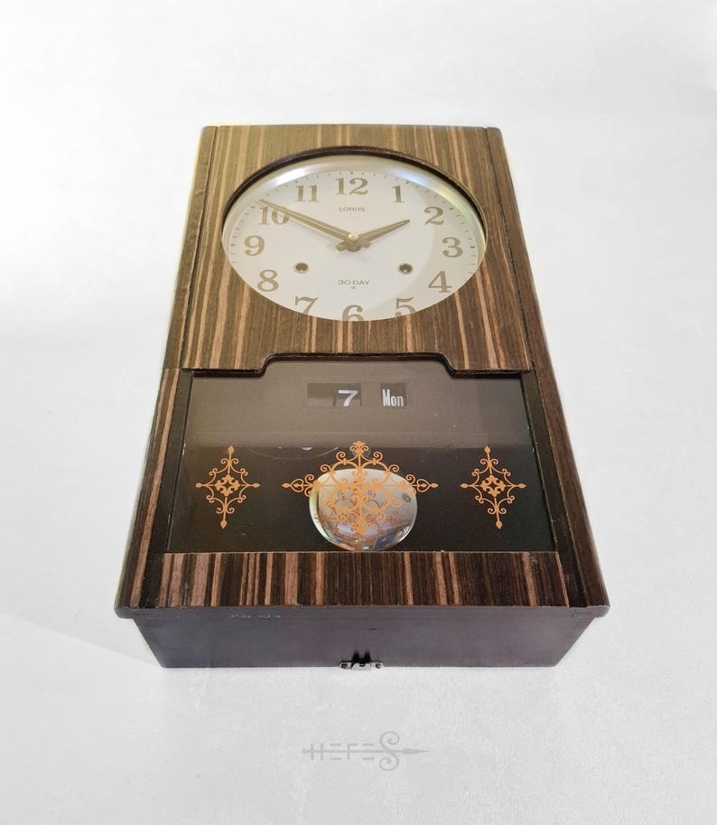 May include: A vintage Lorus clock with a wooden frame and a round clock face. The clock face has black numerals and hands. Below the clock face is a display showing the date and day. The clock has decorative gold accents.