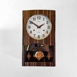 May include: A rectangular wooden clock with a round clock face. The clock face is white with black numerals and hands. The clock has a date display and decorative gold accents. The brand name "LORUS" is visible.