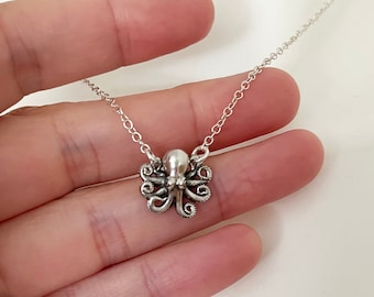 Puede incluir: Collar con colgante de pulpo plateado. El pulpo tiene tentáculos detallados y una cabeza redonda. La cadena del collar es delicada y plateada. El colgante mide aproximadamente 2,5 cm de ancho.