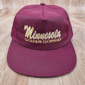 May include: A maroon baseball cap with a flat peak. The cap displays the word "Minnesota" in gold script, arched across the front, with "GOLDEN GOPHERS" below. The cap is made of a sturdy fabric.