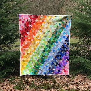 May include: A large rainbow-coloured quilt made of many different fabric hexagons. The quilt is hanging on a clothesline in a woodland setting.