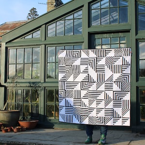 Puede incluir: Una colcha geométrica en blanco y negro con un patrón de rayas y triángulos. La colcha se sostiene frente a un edificio de vidrio verde.