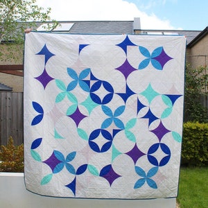 Könnte beinhalten: Eine weiße Steppdecke mit einem geometrischen Muster aus blauen, türkisfarbenen und violetten Kreisen und Rauten. Die Steppdecke hängt an einer Wäscheleine in einem Garten.