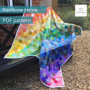 Può includere: Una trapunta con un gradiente arcobaleno di piccoli pezzi di tessuto esagonali disposti in uno spettro dal rosso, arancione e giallo al verde, blu, indaco e viola. La trapunta ha un bordo bianco e include il testo Rainbow Hexie PDF pattern e Cait Lisle Designs.