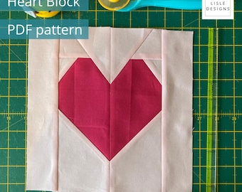 May include: A square fabric quilt block featuring a central dark pink heart shape surrounded by light pink fabric panels. The block is positioned on a green cutting mat with a yellow rotary cutter, a pin cushion, and a clear ruler showing inch markings.