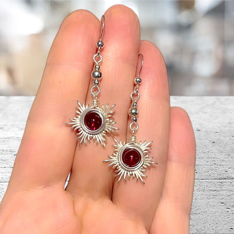 May include: A pair of silver earrings with a red gemstone in the center of each earring. The earrings are shaped like a sun with rays extending out from the center.