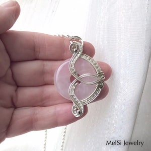 May include: A silver wire wrapped pendant featuring a light pink rose quartz stone. The pendant is shaped like a figure eight and hangs from a silver chain.