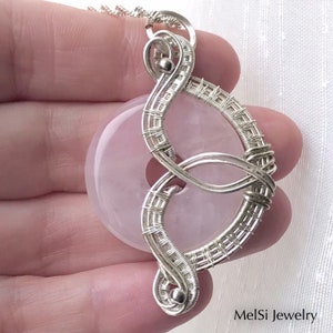 May include: A silver wire wrapped pendant featuring a pale pink, round gemstone. The pendant is shaped like a figure eight and has a delicate, intricate design. The pendant is hanging from a silver chain.