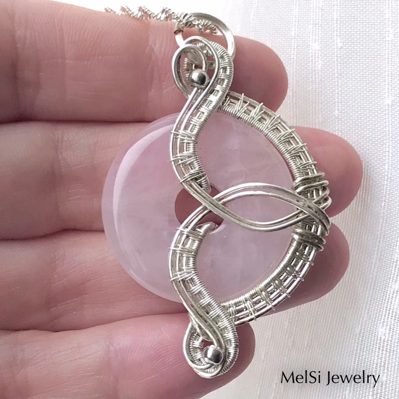 May include: A silver wire wrapped pendant featuring a pale pink, round gemstone. The pendant is shaped like a figure eight and has a delicate, intricate design. The pendant is hanging from a silver chain.