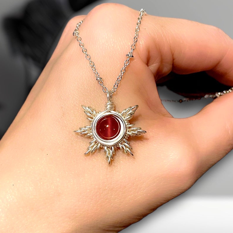 May include: A silver chain necklace with a red gemstone sun pendant. The sun is made of wire and has a red gemstone in the center.