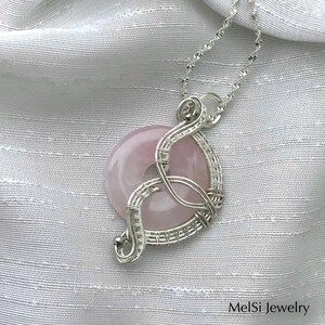 May include: A silver wire wrapped pendant featuring a light pink rose quartz stone. The pendant is hanging from a silver chain on a white fabric background. MelSi Jewelry is printed in the bottom right corner of the image.