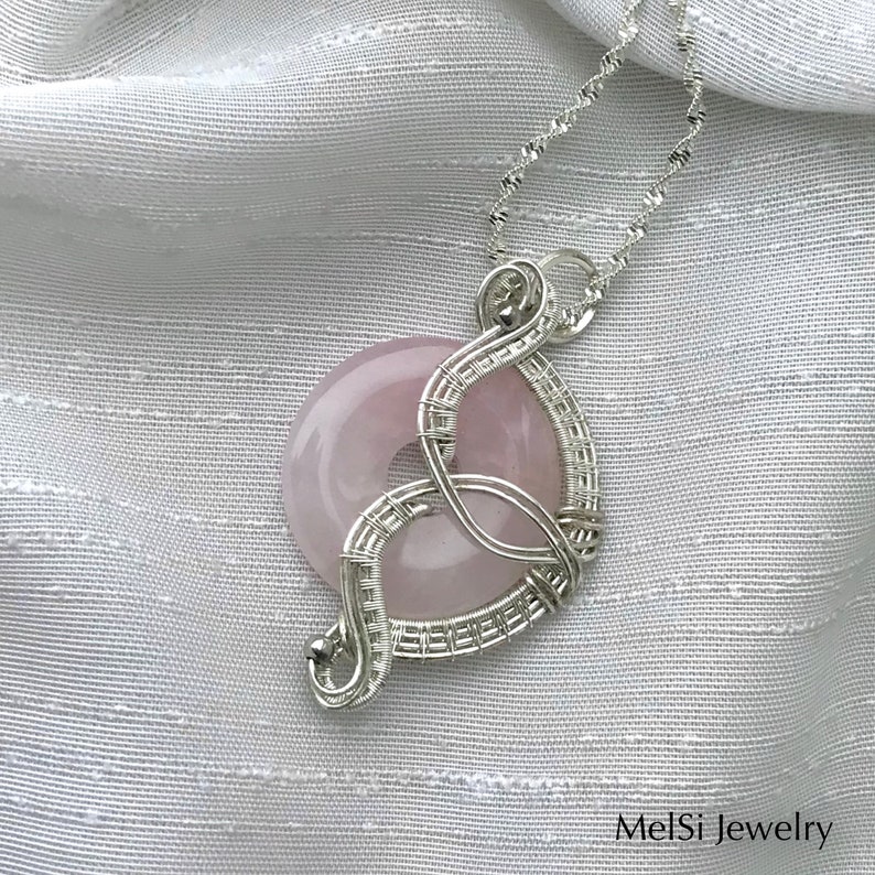 May include: A silver wire wrapped pendant featuring a light pink rose quartz stone. The pendant is hanging from a silver chain on a white fabric background. MelSi Jewelry is printed in the bottom right corner of the image.