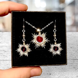 May include: A silver necklace and earrings set with red gemstone sunburst pendants. The necklace has a delicate chain and the earrings have small silver beads.