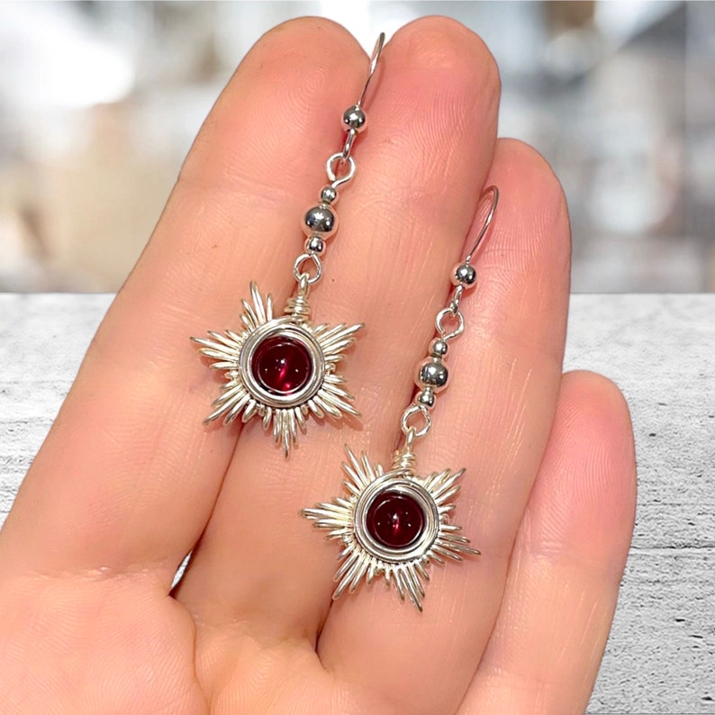 May include: Silver earrings with a red gemstone in the center of a starburst design. The earrings have a chain of silver beads hanging from the starburst.