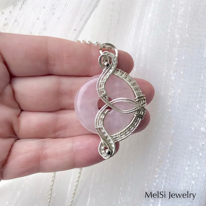 May include: A silver wire wrapped pendant featuring a pink rose quartz stone. The pendant is shaped like a figure eight and hangs from a silver chain.