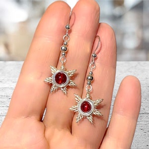 May include: Silver dangle earrings with a red gemstone in the center of a wire-wrapped star design. The earrings have a silver bead chain.