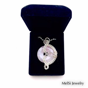 May include: A silver wire wrapped pendant featuring a pink rose quartz stone. The pendant is displayed in a blue velvet jewelry box. MelSi Jewelry is printed on the bottom of the image.