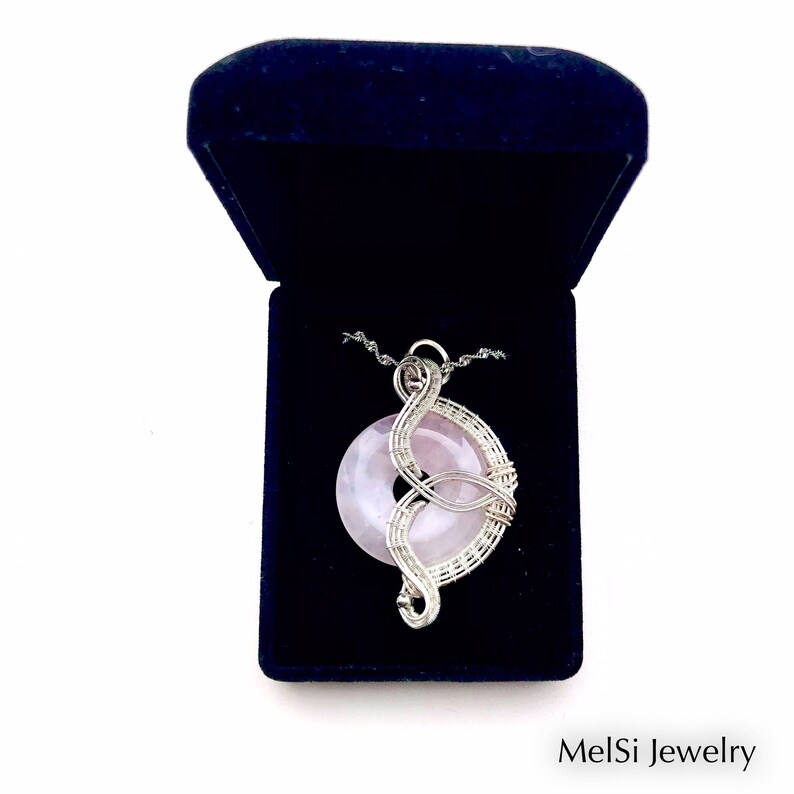 May include: A silver wire wrapped pendant featuring a pink rose quartz stone. The pendant is displayed in a blue velvet jewelry box. MelSi Jewelry is printed on the bottom of the image.