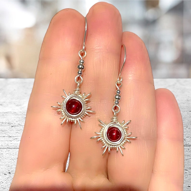 May include: Silver dangle earrings with a red gemstone in the center of a wire-wrapped sun design.
