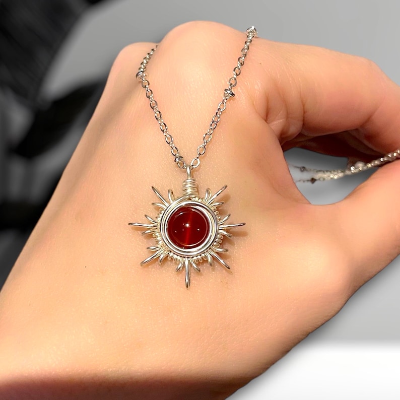 May include: A silver necklace with a red carnelian stone set in a wire-wrapped sun pendant.