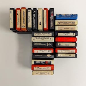 May include: A collection of vintage 8-track tapes in various colors, including red, orange, black, and blue. The tapes are arranged in a black display rack, showcasing album titles and artist names. The collection includes titles like "Country Teardrops" and "A Star is Born."