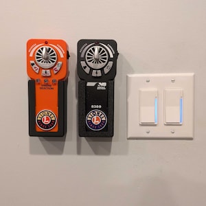 May include: Two black and orange Lionel train controllers mounted on a white wall. The controllers have a black dial with a silver knob and a black button labeled "Shift". The controllers have a black and white Lionel logo with the text "Lionel Since 1900".