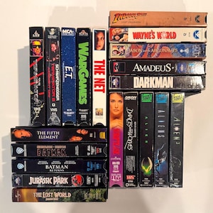 May include: A collection of VHS tapes featuring movie titles like "Terminator 2," "E.T.," "Wargames," and "Jurassic Park." The tapes are stacked vertically, showcasing their colorful labels and movie artwork. A nostalgic display of classic films.