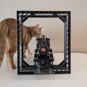 May include: A ginger cat is looking at a black toy train engine with a red number 653 on the side. The train is inside a black plastic frame.