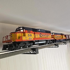 May include: A model train locomotive painted in yellow, orange, and black with the text C&O 8204 and Chessie System printed on the side. The train is positioned on a raised track section supported by black metal trestle supports against a wall.