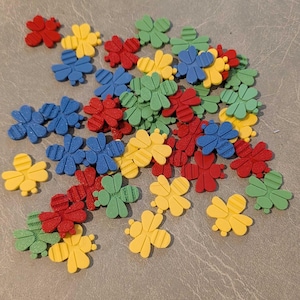 May include: A collection of colorful, flower-shaped embellishments in red, yellow, blue, and green. The plastic flowers have a textured surface and are scattered across a neutral background. These decorative elements could be used for crafts or other creative projects.