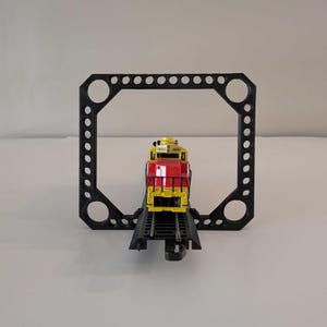 May include: A yellow and red toy train locomotive with black wheels and a black frame is on a black track. The train is going through a black plastic frame with holes.