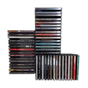 May include: A collection of CDs stacked in three groups. The CDs have various album titles and artist names visible on the covers. The cases are mostly black with some red and white accents. The image is on a white background.