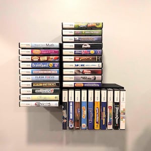 May include: A collection of Nintendo DS video game cartridges stacked on black shelves. The game cases are white and black, with colorful game titles visible, including "BrainQuest," "Deal or No Deal," and "High School Musical."