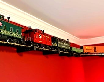 May include: A model train set displayed on a wall-mounted shelf, featuring a green locomotive, a red caboose, a dark green passenger car, and an orange freight car. The train cars are positioned in a line along the track against a red wall.