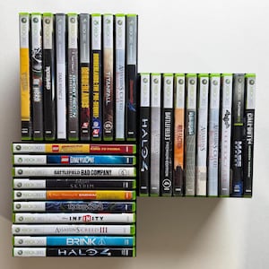 May include: A collection of Xbox 360 video game cases arranged in two vertical stacks and one horizontal stack. The spines display various titles including Halo 4, Assassin's Creed, Borderlands, Mass Effect, and Call of Duty, featuring diverse colors.