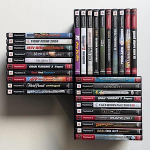 May include: A collection of PlayStation 2 video game cases arranged in three stacks. The titles include Bass Fishing Duel, Fight Night 2004, ATV Offroad Fury 2, Kessen II, Final Fantasy X-2, Gran Turismo 3, Seek and Destroy, Trivial Pursuit, and Wreckless.