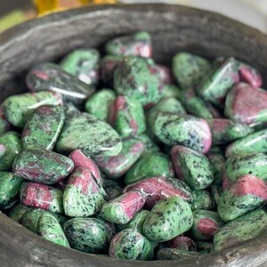 May include: A bowl filled with green and pink tumbled stones. The bowl is made of dark brown wood and has a rough texture. The stones are a mix of green and pink, with some having a speckled pattern.