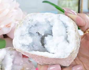 May include: Two white crystal geodes with a rough exterior and a hollow interior filled with white crystals. The geodes are being held in a hand with a gold ring.