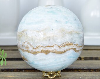May include: A blue and white agate sphere with brown banding sits on a gold metal stand. The sphere is on a wooden surface.