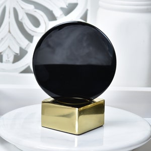 Peut inclure: Un objet décoratif noir et or avec un dessus rond noir et une base carrée dorée. L'objet est posé sur une assiette blanche.