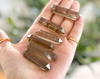 Pode incluir: Seis pontas de cristal de quartzo fumado, também conhecidas como quartzo marrom, estão dispostas em uma fileira em uma mão. As pontas são de tamanhos e formas diferentes, mas todas têm um tom marrom semelhante.