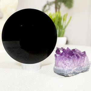 Op de afbeelding: Een zwart, cirkelvormig object op een witte standaard, naast een cluster van paarse amethistkristallen. De kristallen hebben een kleurovergang van lichtpaars naar een grijze basis. Een wit beeld en een potplant staan op de achtergrond.