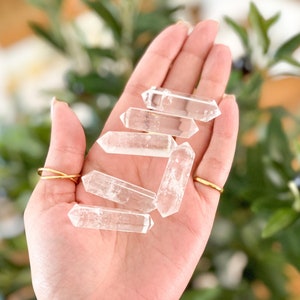 May include: A collection of six clear quartz crystal points, also known as clear quartz wands, are held in a hand. The points are smooth and polished, with a slightly pointed tip.