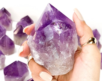 Pode incluir: Uma grande ponta de cristal de ametista com uma base clara e translúcida e um topo roxo escuro. O cristal está sendo segurado em uma mão com um anel de ouro. Há outros cristais de ametista no fundo.