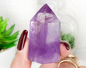 Pode incluir: Uma ponta de cristal de ametista roxa, também conhecida como cristal de terminação única, é uma gema natural com uma ponta pontiaguda. O cristal é segurado na mão com um anel no dedo.