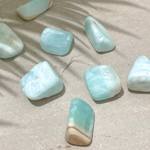 May include: A collection of eight smooth, light blue aquamarine tumbled stones. The stones are various shapes and sizes, and some have white and brown markings.