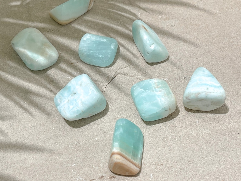 May include: A collection of eight smooth, light blue aquamarine tumbled stones. The stones are various shapes and sizes, and some have white and brown markings.