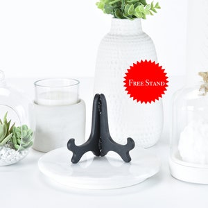 May include: A white ceramic vase with a textured surface, filled with green plants. A black metal display stand is on a white ceramic plate. A glass cloche with a white object inside is on a white ceramic base. A glass cloche with a small succulent plant inside is on a white ceramic base. The text "Free Stand" is on a red starburst.