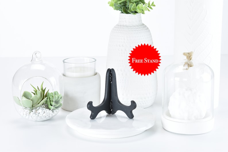 May include: A white ceramic vase with a textured surface, filled with green plants. A black metal display stand is on a white ceramic plate. A glass cloche with a white object inside is on a white ceramic base. A glass cloche with a small succulent plant inside is on a white ceramic base. The text "Free Stand" is on a red starburst.